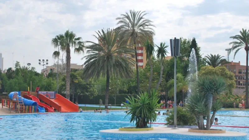 Piscina del Parque de Benicalap en Valencia con toboganes y zonas verdes, una de las mejores piscinas de Valencia para verano Vista general de la piscina del Parque de Benicalap en Valencia, con toboganes, palmeras y zonas ajardinadas